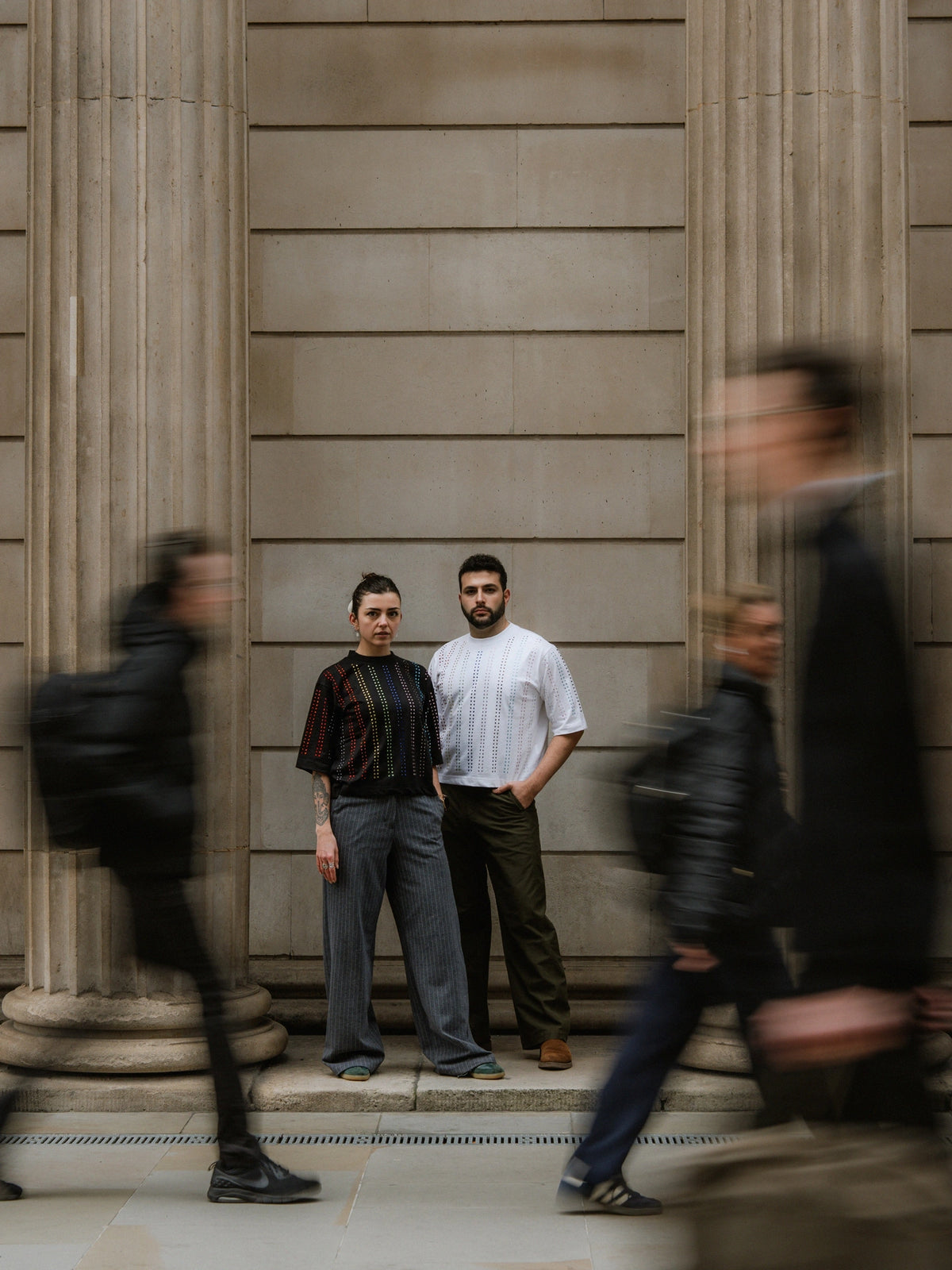 Two models standing between classical columns wearing Haus of Mx. Proud T-shirts with rainbow crystal stripes pride inspired, while blurred pedestrians walk past in motion.