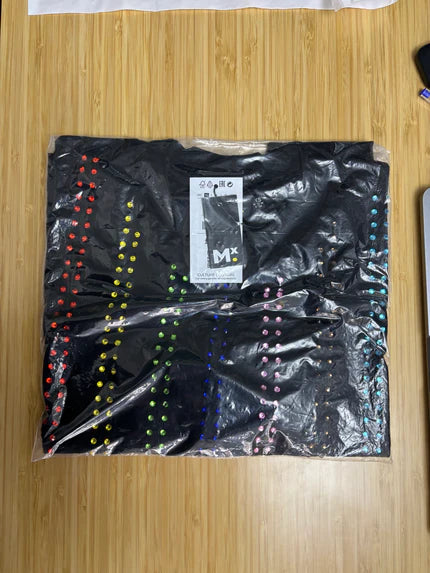 Folded black Haus of Mx. T-shirt in clear polybag with vertical rainbow rhinestone stripes,  pride inspired, on a wooden table