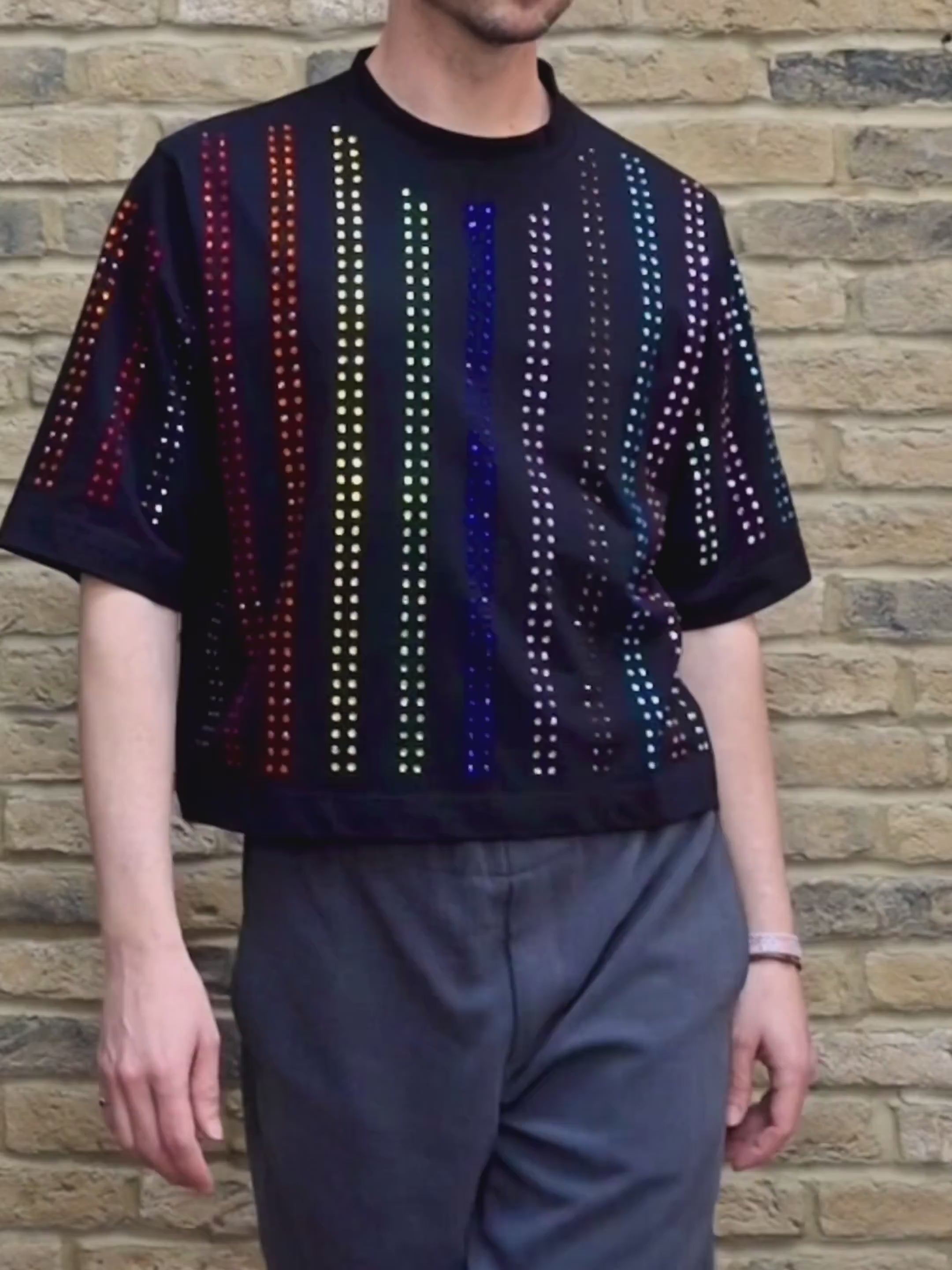 Model wearing black Proud T-shirt with rainbow crystal lines, pride inspired, against a brick wall